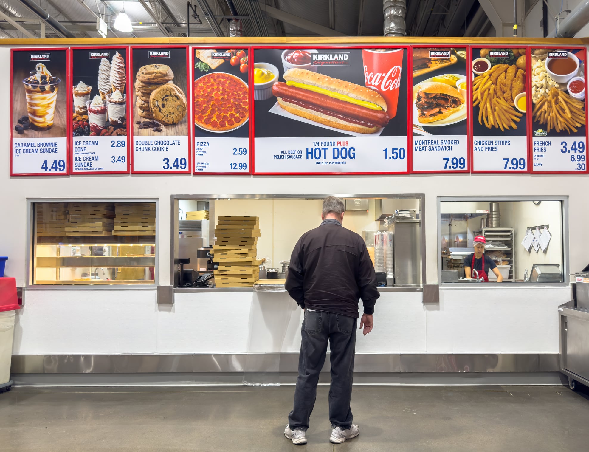 A man stands in front of a Costco food court counter beneath a large menu board listing items and prices.
