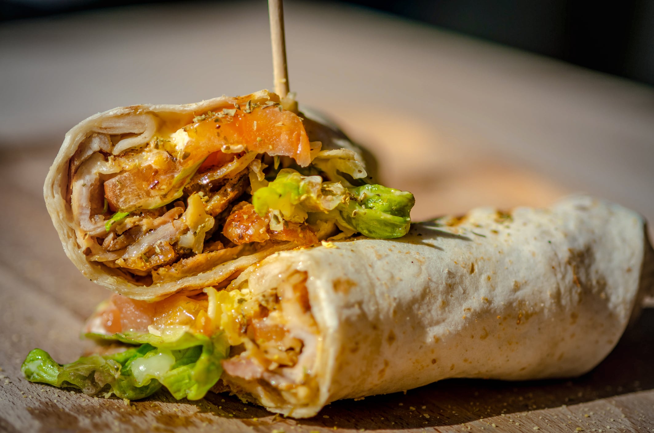 Two halves of a chicken wrap with lettuce, tomato, and sauce, held together with a wooden pick on a wooden board.