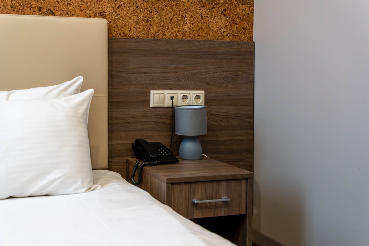 A bedside table with a landline phone and a small gray lamp, situated next to a bed with white pillows.