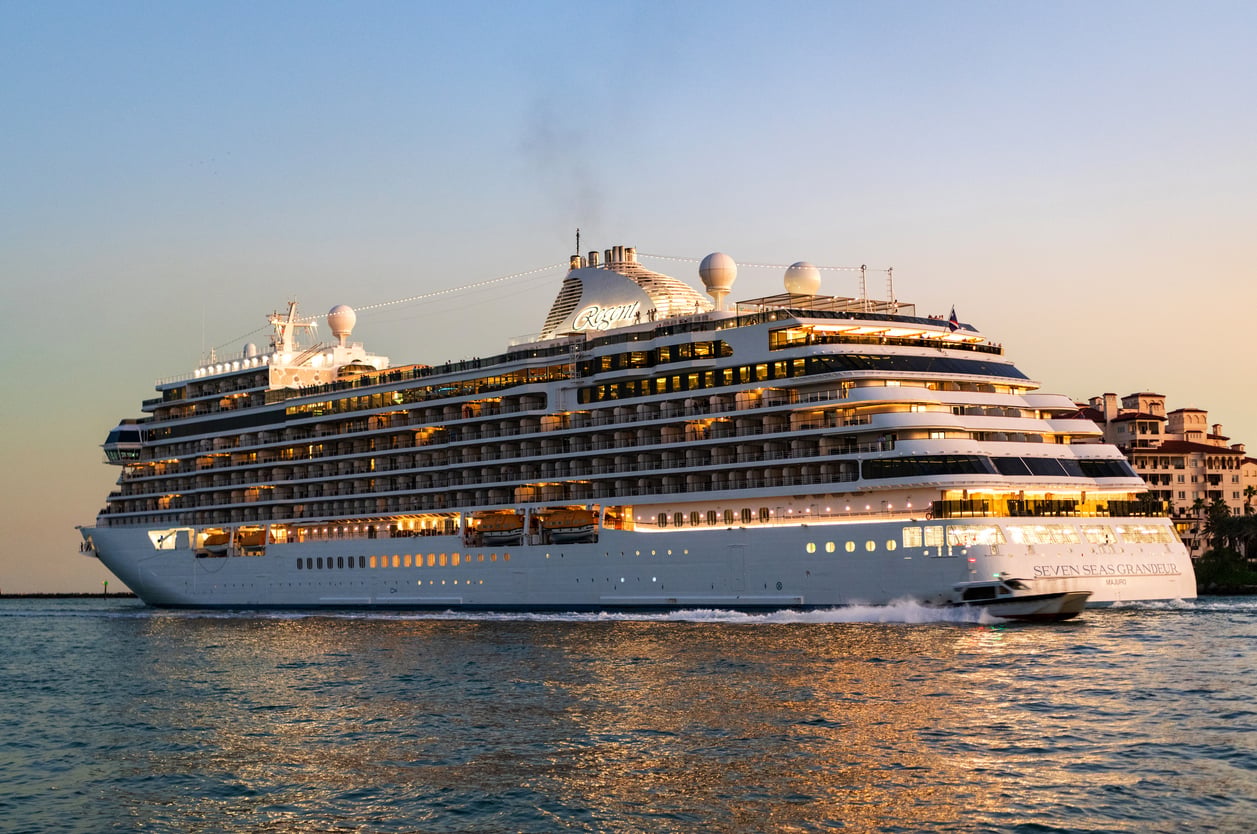 The Seven Seas Grandeur cruise ship, a hub for cruise entertainment, sails on the water at sunset with a small boat nearby.