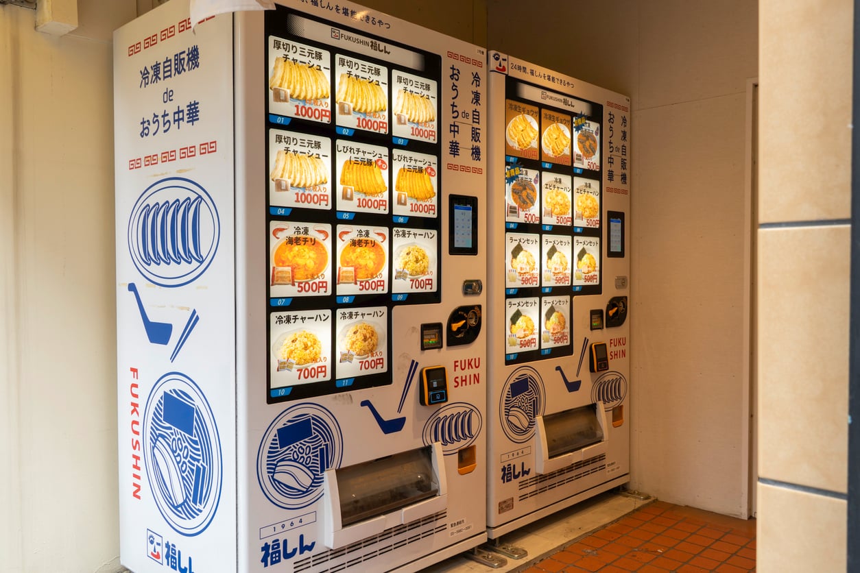 Two white vending machines selling frozen Chinese meals, featuring menu photos with prices and blue line-art illustrations of bowls and spoons.
