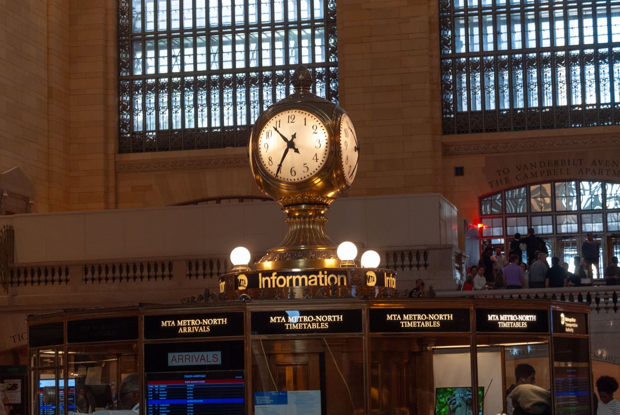 The four-faced brass clock atop the information kiosk in Grand Central Terminal, with digital arrival and timetable screens visible below.
