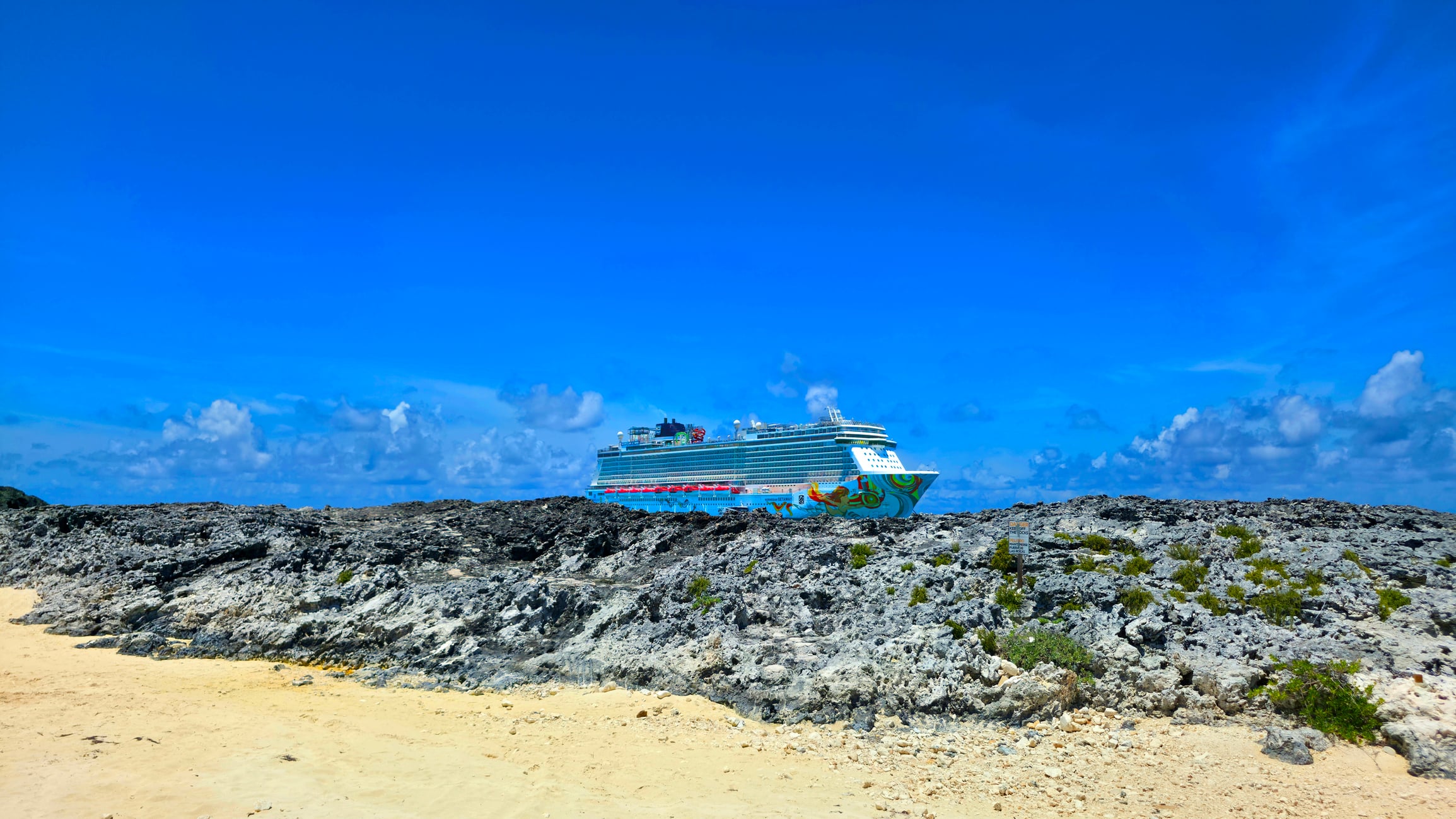 A large cruise ship anchored behind a rocky shoreline under a bright blue sky.
