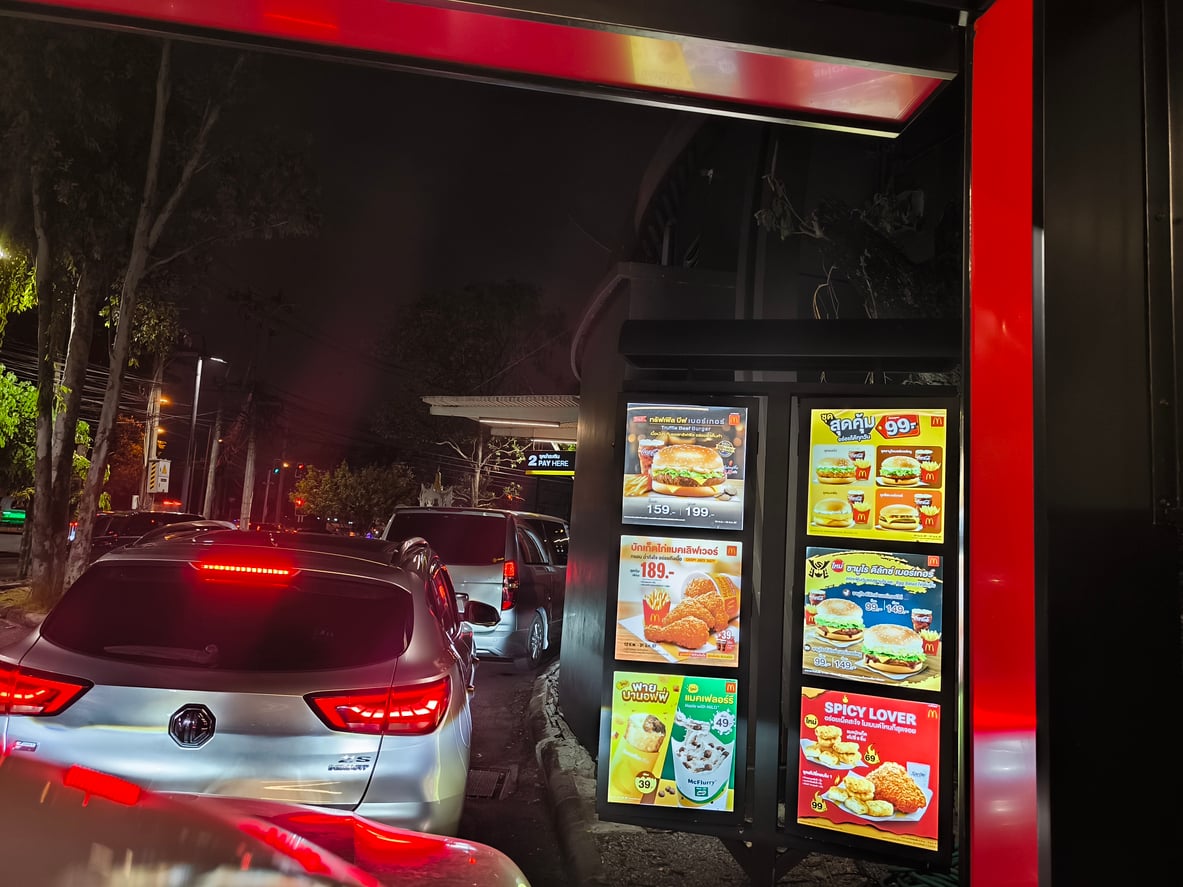 Cars waiting in a McDonald’s drive-thru line at night next to a digital menu board displaying meal options and prices.
