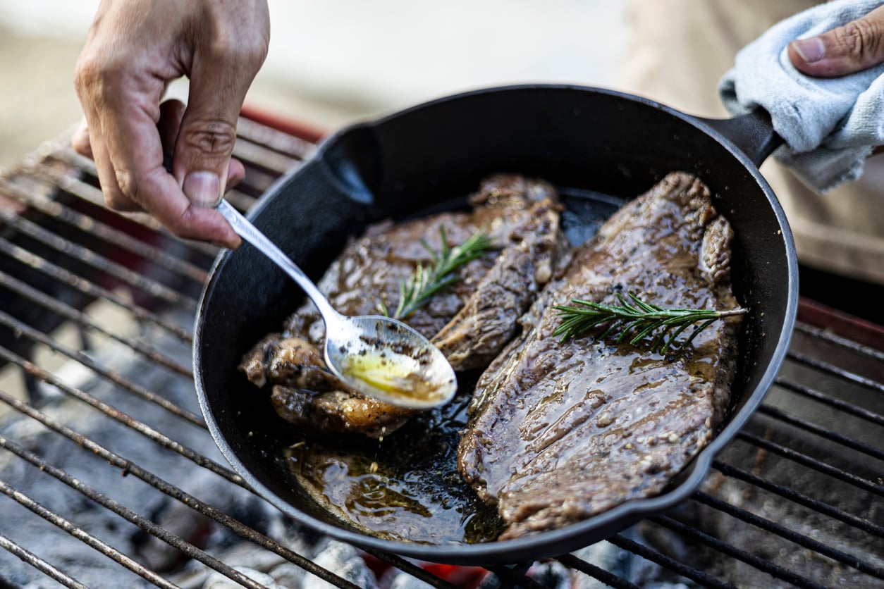 A hand uses a spoon to drizzle butter over two steaks topped with rosemary sprigs in a cast-iron skillet on a grill.