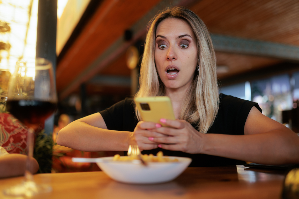 A woman with a surprised expression looks at her yellow smartphone while sitting at a restaurant table with a bowl of food and a glass of wine.