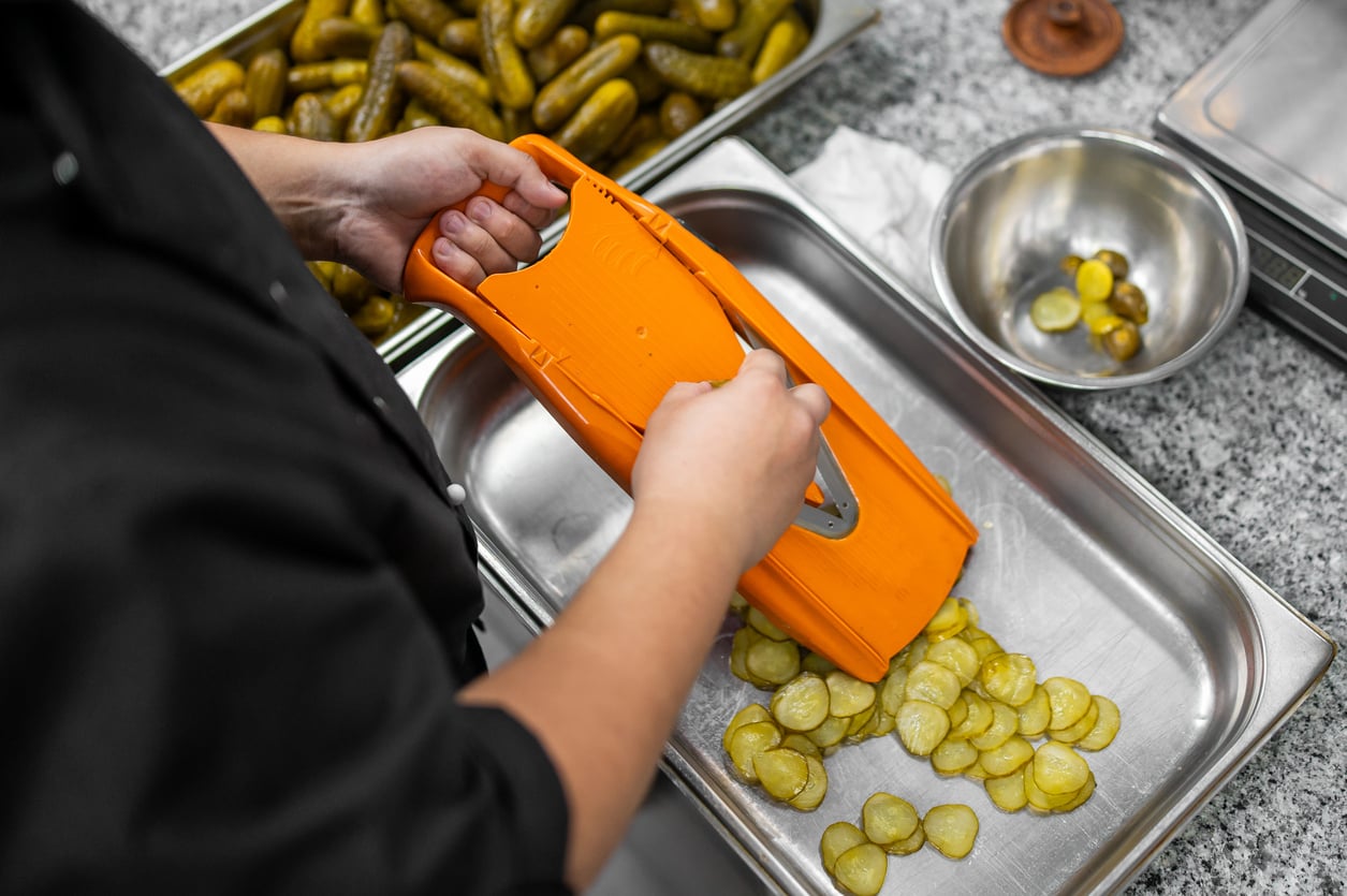 A person in a black uniform uses an orange mandoline to slice pickles into a metal tray.