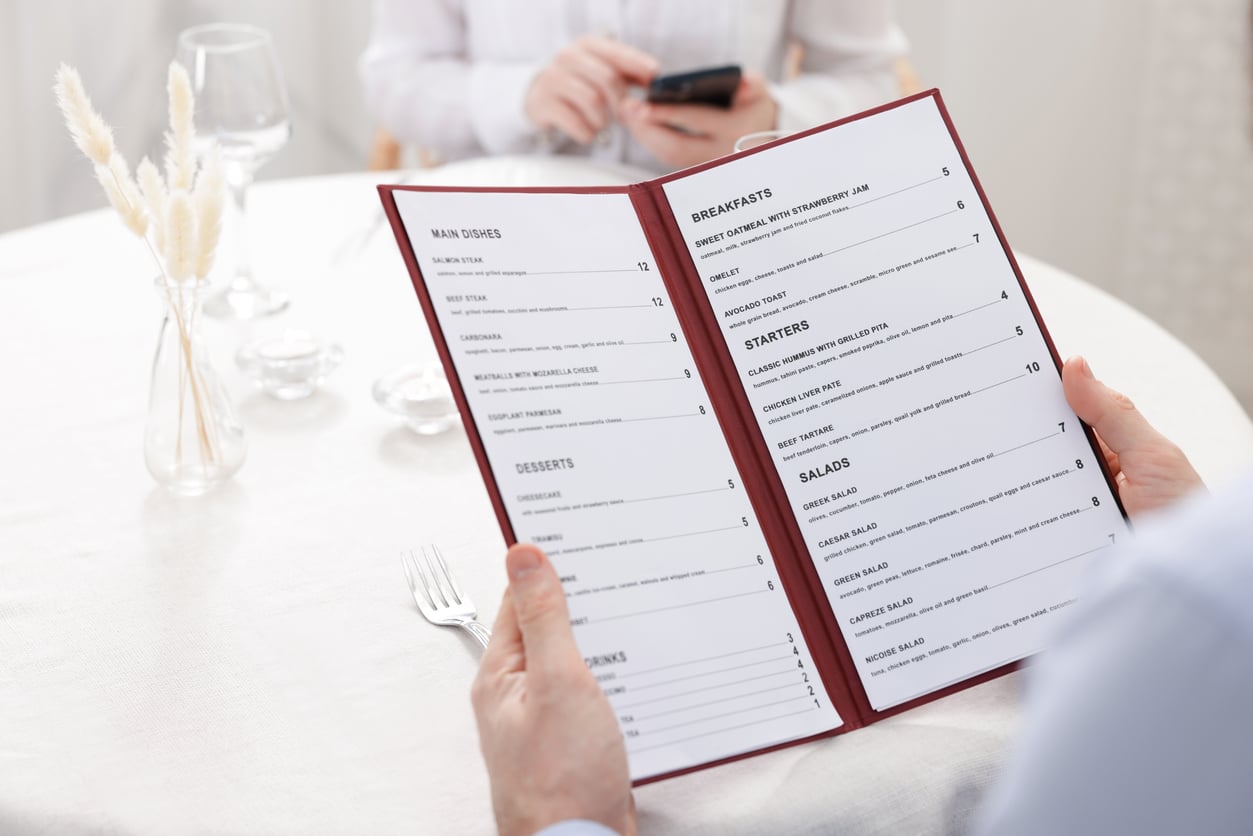 A person holds an open restaurant menu at a table, highlighting different menu tips for main dishes, breakfasts, starters, and salads.