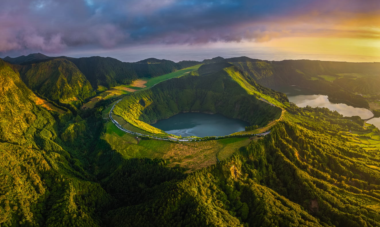 Aerial view of a green volcanic crater lake surrounded by lush, hilly landscape at sunset.