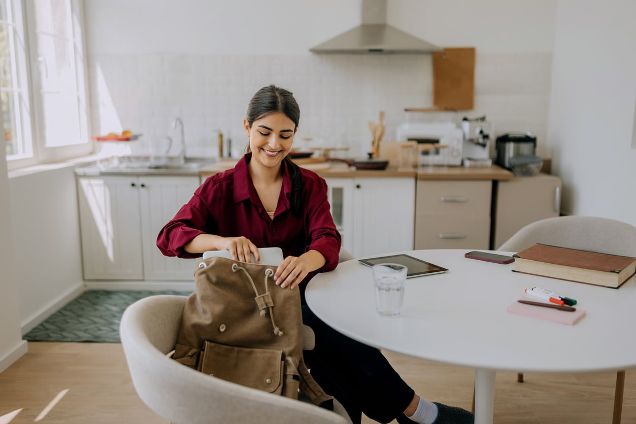 A smiling woman in a maroon shirt packs a laptop into her brown backpack at a kitchen table.