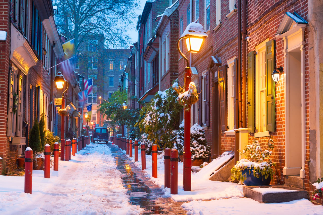 A snowy cobblestone street lined with historic brick houses and glowing vintage streetlights at dusk.