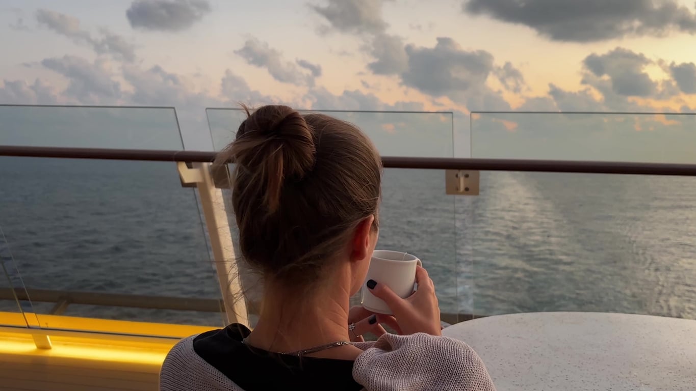 A woman seen from behind stands on a cruise ship balcony at sunset, holding a white mug while looking out at the ocean.