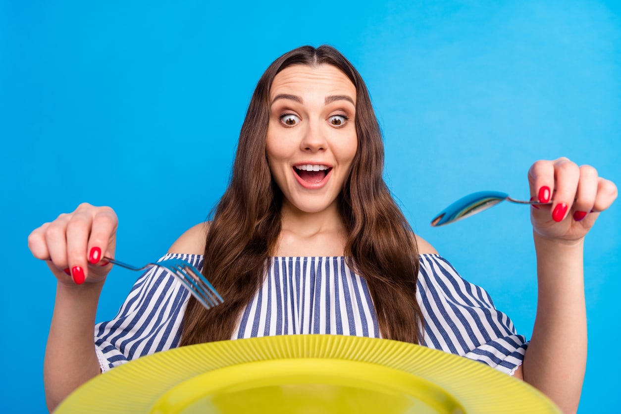 A woman with an excited expression holds a fork and spoon over an empty yellow plate.