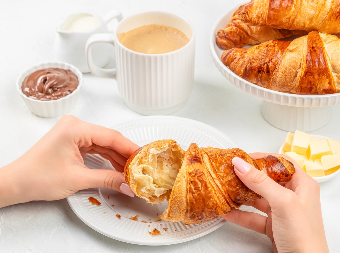Hands pulling apart a flaky croissant on a white plate, surrounded by a cup of coffee, chocolate spread, butter, and a tiered stand of croissants.