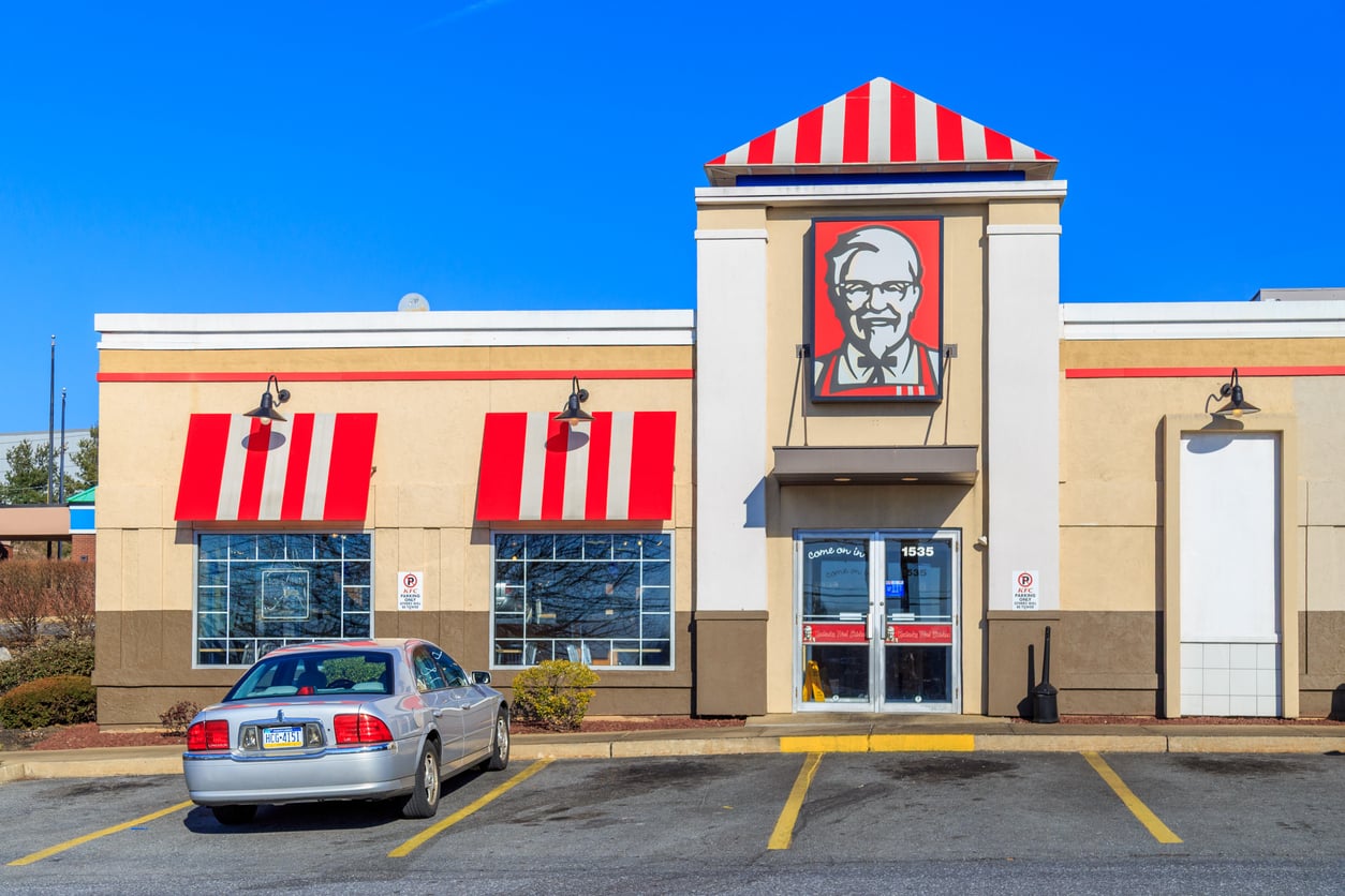 A KFC restaurant building with red and white striped awnings and a large Colonel Sanders sign above the entrance, with a silver sedan parked in front.