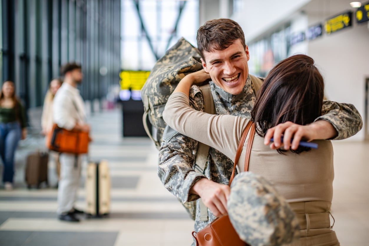 A smiling soldier in camouflage uniform embraces a woman in an airport terminal.