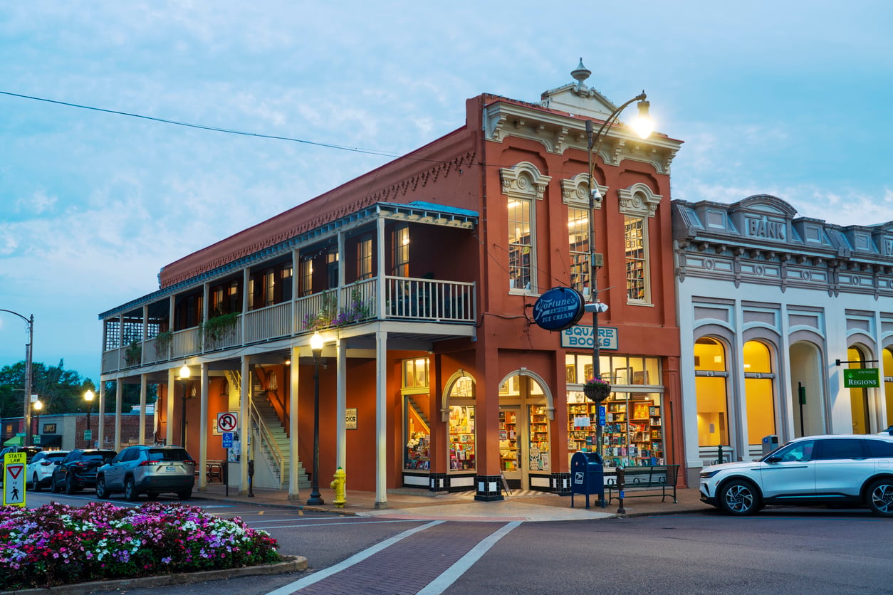 A two-story red brick building on a street corner housing Square Books and Fortune’s Ice Cream, adjacent to a historic bank building at twilight.