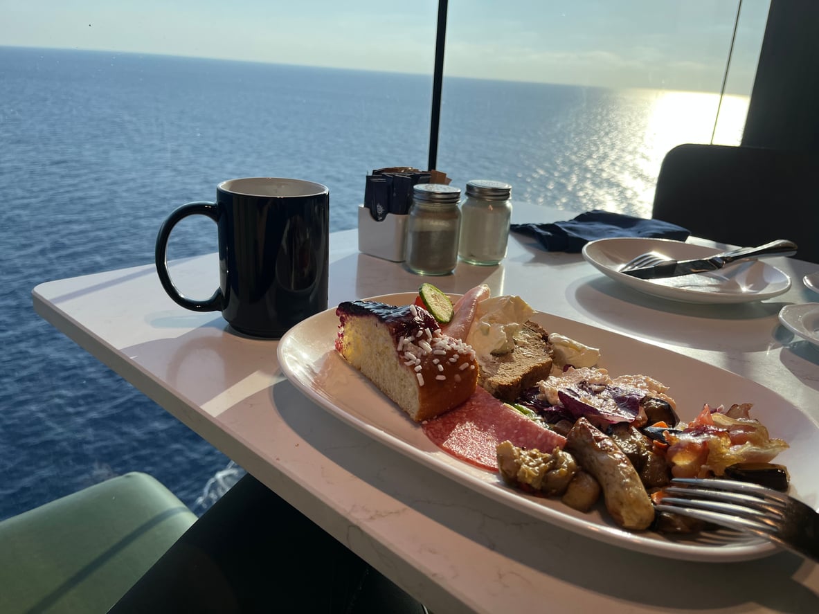 Cruise Ship Dining Tricks:
A breakfast plate featuring a slice of cake, deli meat, bread, and roasted vegetables, set on a table next to a black mug with the ocean in the background.