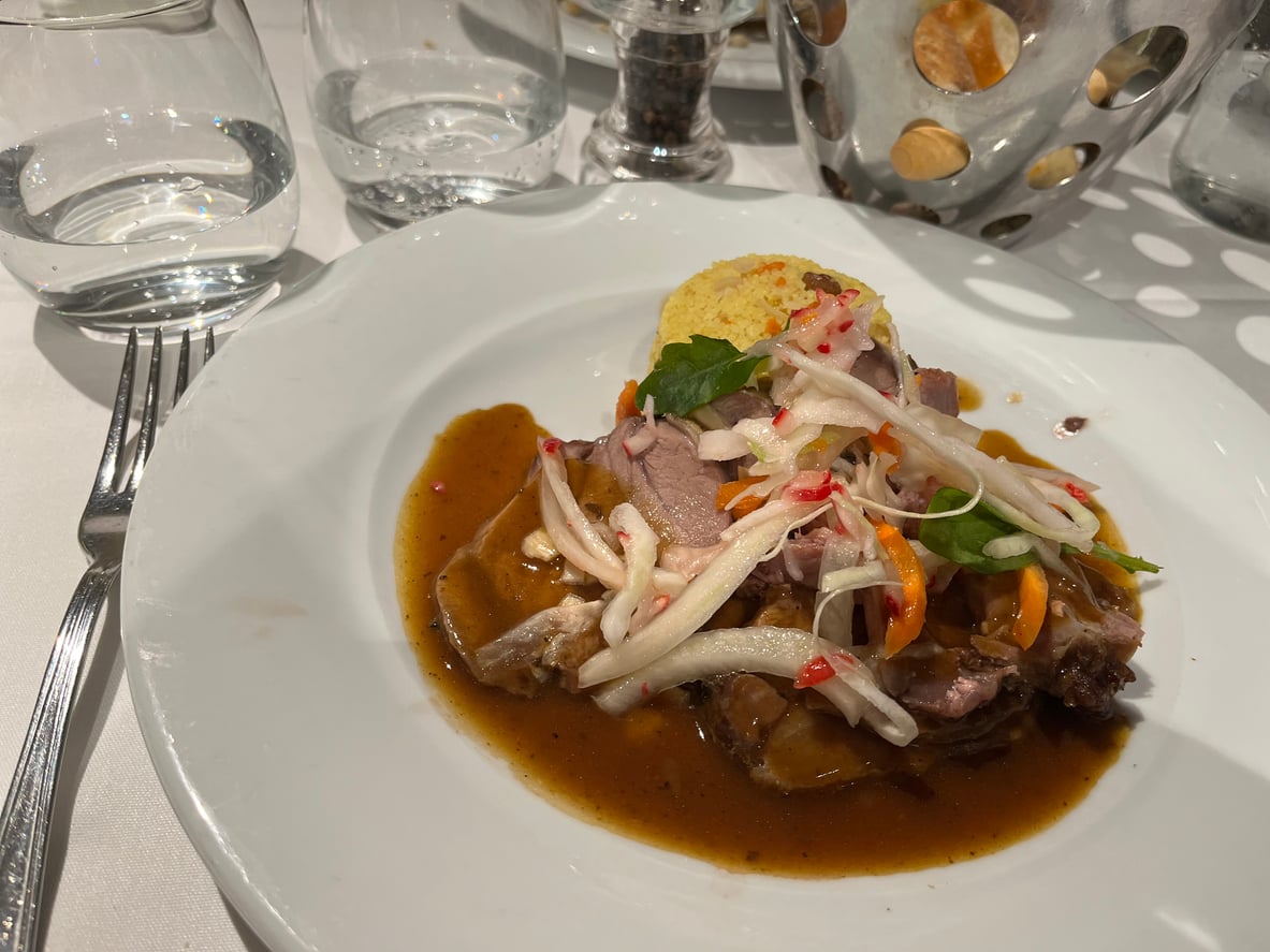 A plated dish of braised meat in gravy topped with a slaw of shredded vegetables and herbs, served with a side of yellow grains.