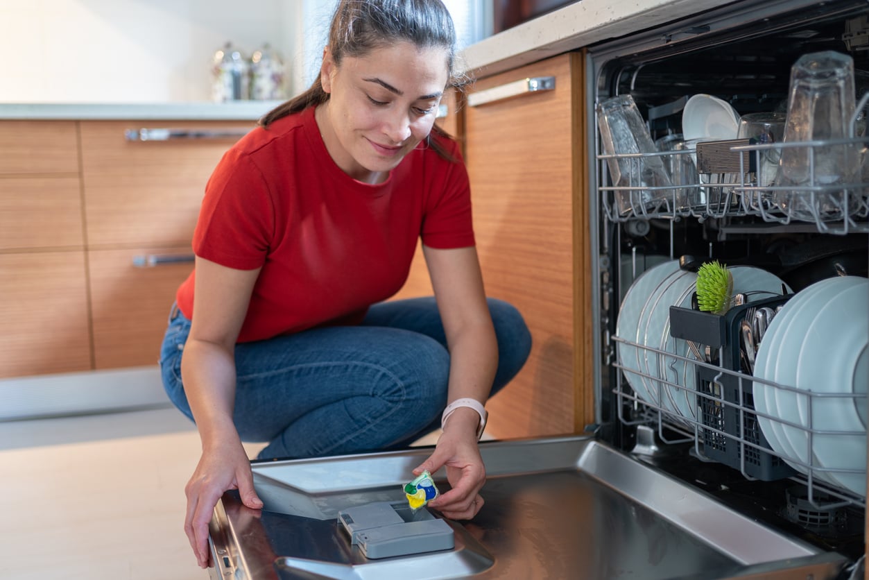 A woman in a red shirt places a dishwasher detergent pod into the soap dispenser of an open dishwasher.