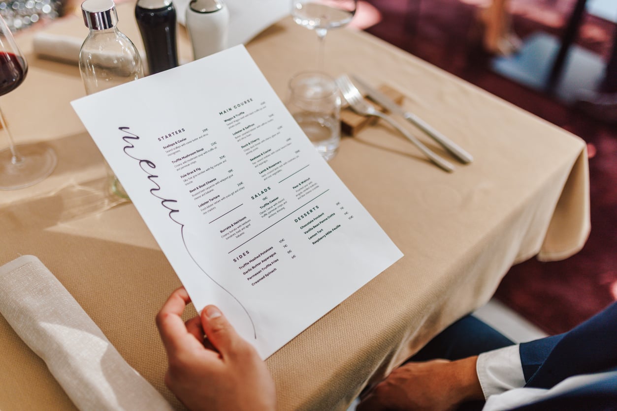 A person holds a restaurant menu with tips on starters, main courses, salads, sides, and desserts over a table set with silverware and glasses.