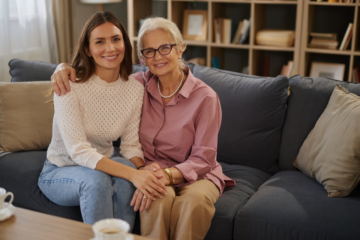 A younger woman and an older woman sitting together on a sofa with their arms around each other, smiling at the camera.