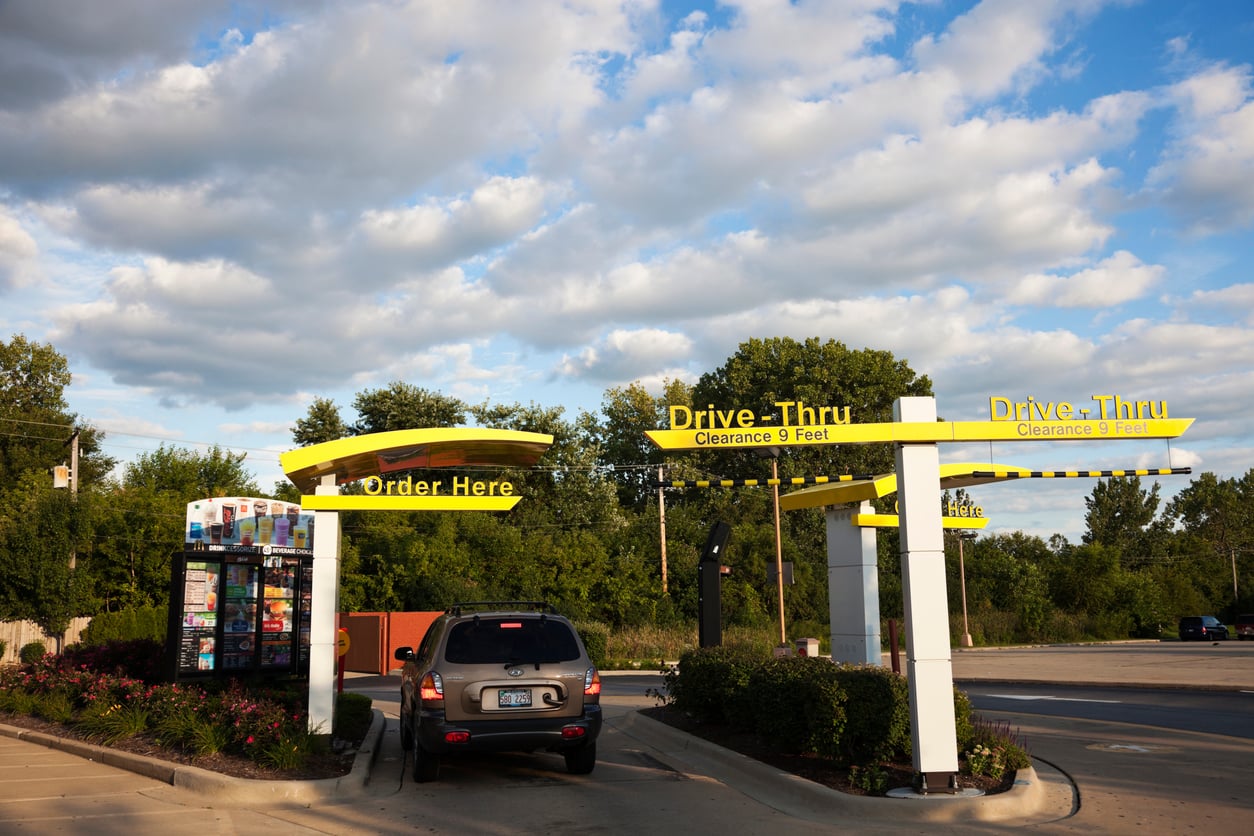 An SUV waits at a fast-food drive-thru lane with yellow “Order Here” and “Drive-Thru” signs.