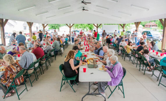 A large group of people eating cheap meals for crowds at long tables under a covered outdoor pavilion.
