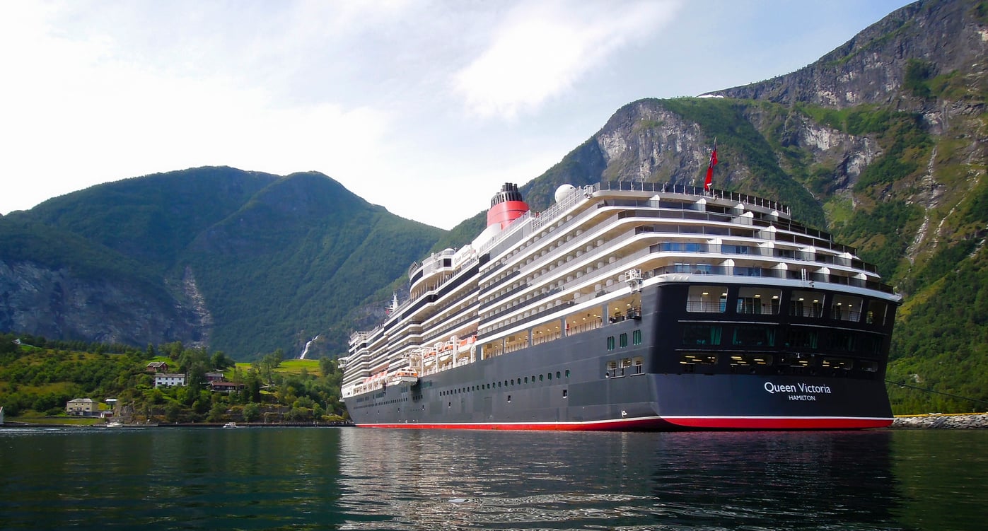 The Queen Victoria cruise ship, known for its cruise entertainment, is docked in a harbor surrounded by steep, green mountains.