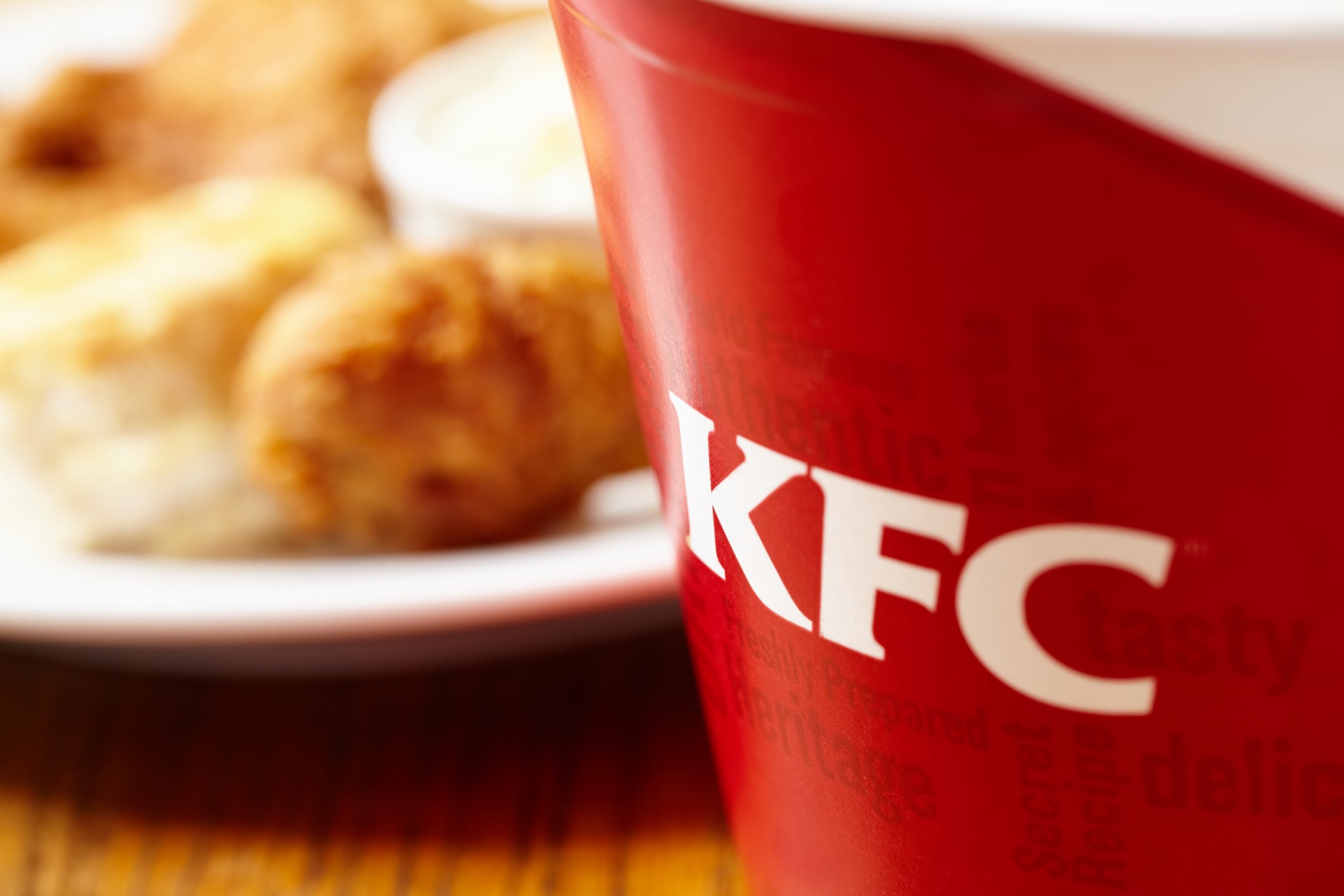 A KFC bucket in the foreground with a blurred plate of fried chicken and a biscuit in the background.