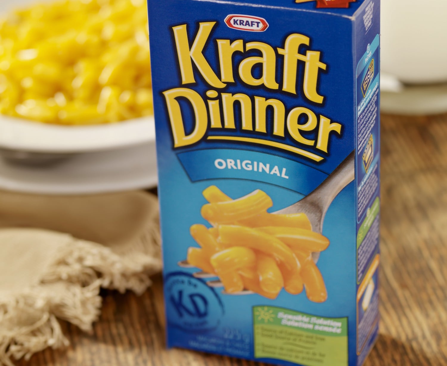 A box of Kraft Dinner Original macaroni and cheese, with a bowl of prepared macaroni and cheese in the background.