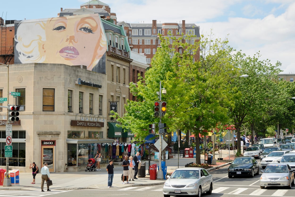 A city street corner featuring a large mural of Marilyn Monroe painted on the side of a building above a Chipotle restaurant, with pedestrians waiting at a crosswalk and cars driving through the intersection.