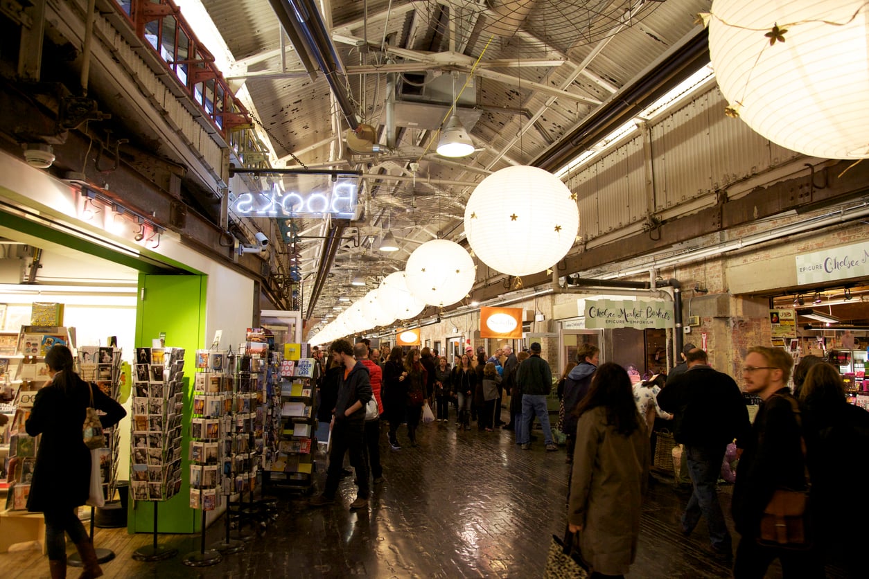 People walking through a long indoor market hall with paper lanterns hanging from the high, industrial ceiling.