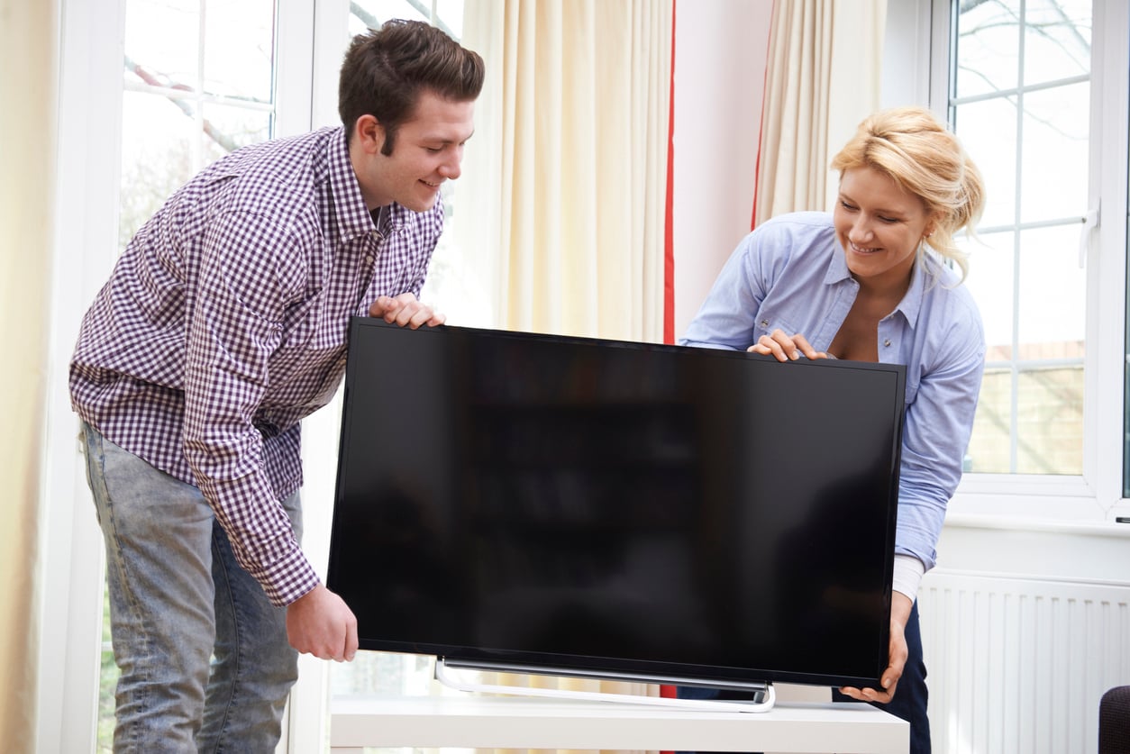A man and a woman smiling as they place a flat-screen television onto a stand.