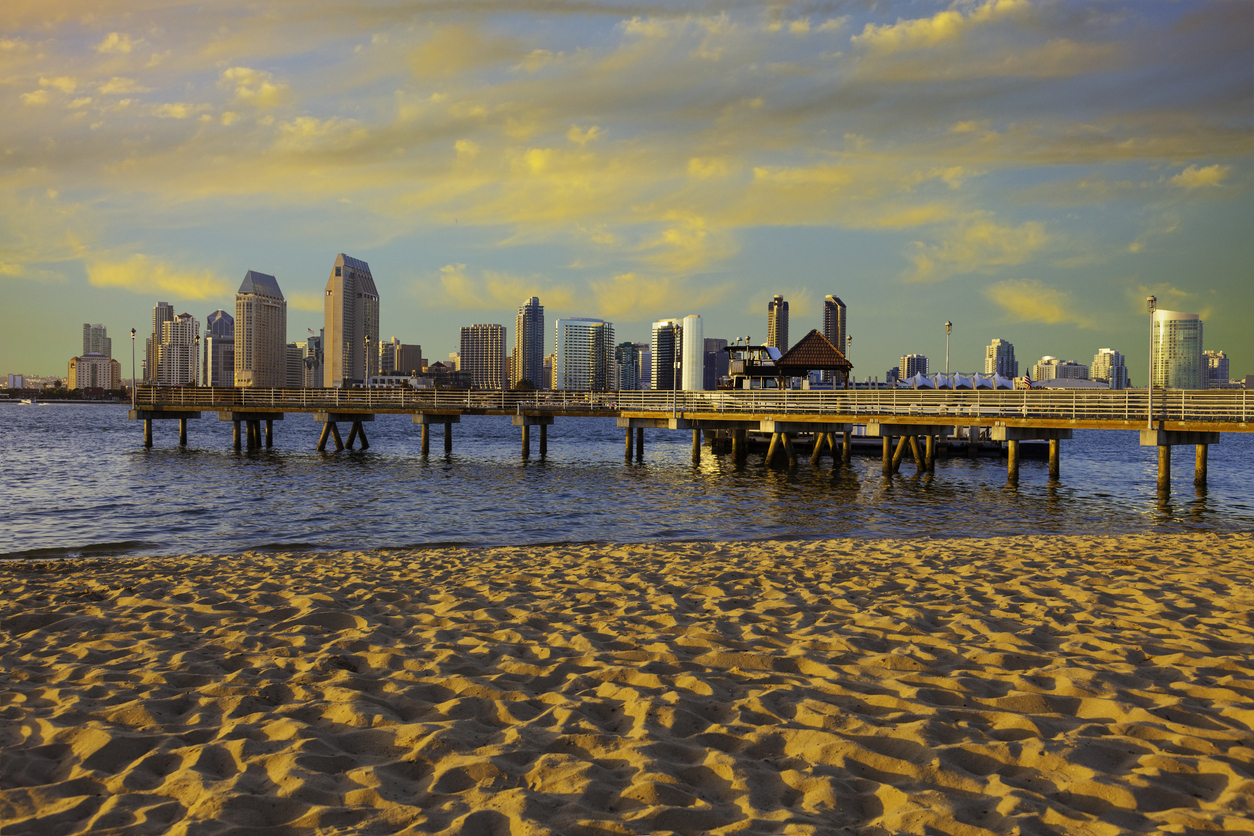 A sandy beach in the foreground leading to a wooden pier that extends into a bay toward the San Diego city skyline at sunset, perfect for trips that feel luxurious.