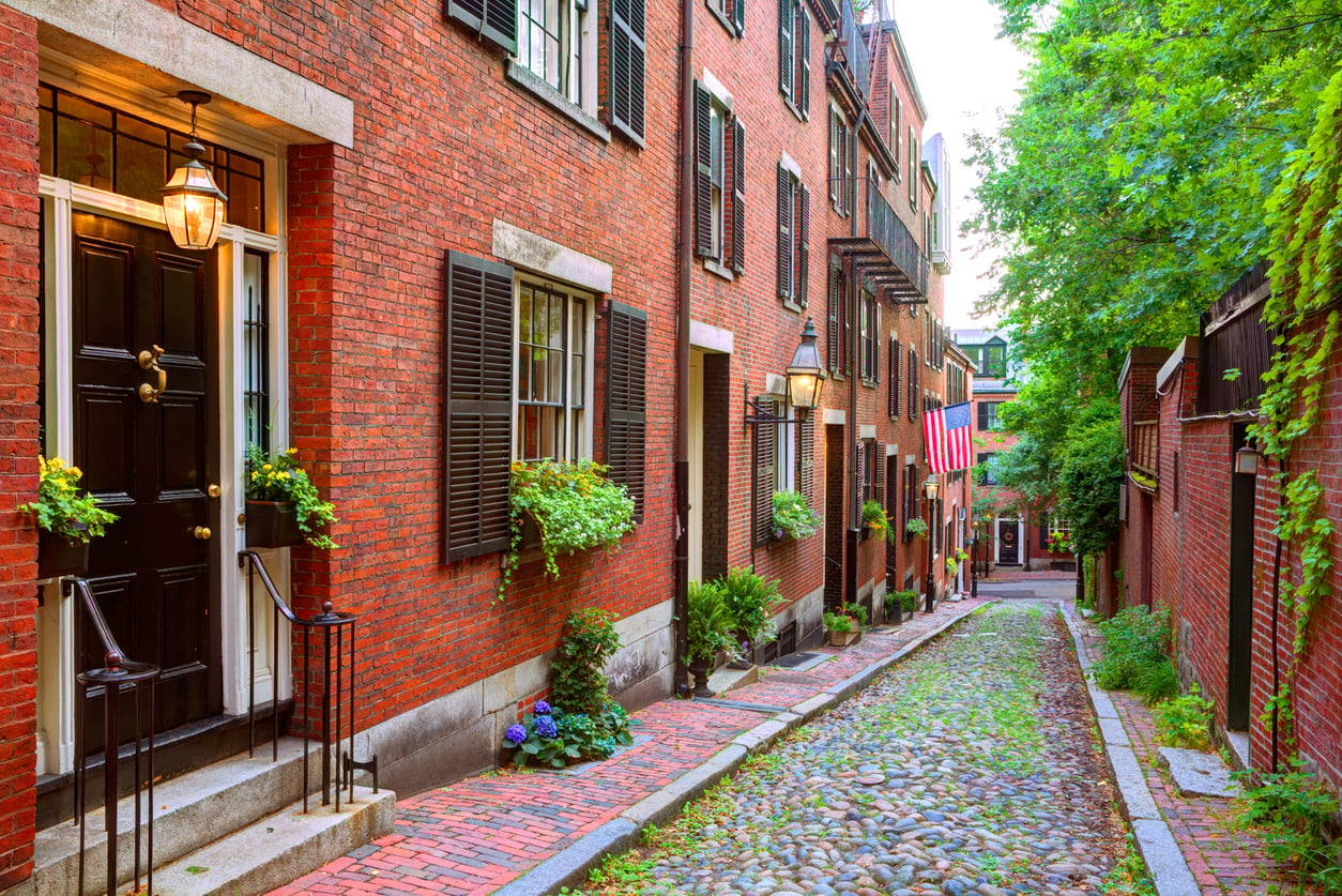 A narrow cobblestone street lined with historic brick row houses featuring black shutters, window boxes with flowers, and an American flag hanging from one of the buildings.