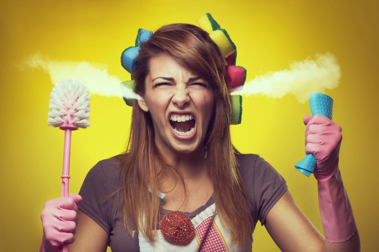 A woman with hair curlers and pink cleaning gloves screams in frustration with steam coming out of her ears, holding a toilet brush and a sponge.