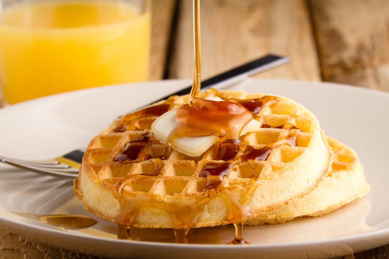Syrup pouring onto a buttered waffle on a plate, with a glass of orange juice in the background.