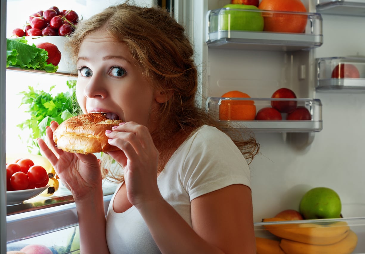 A woman looking surprised while taking a bite of a sandwich in front of an open refrigerator filled with produce.