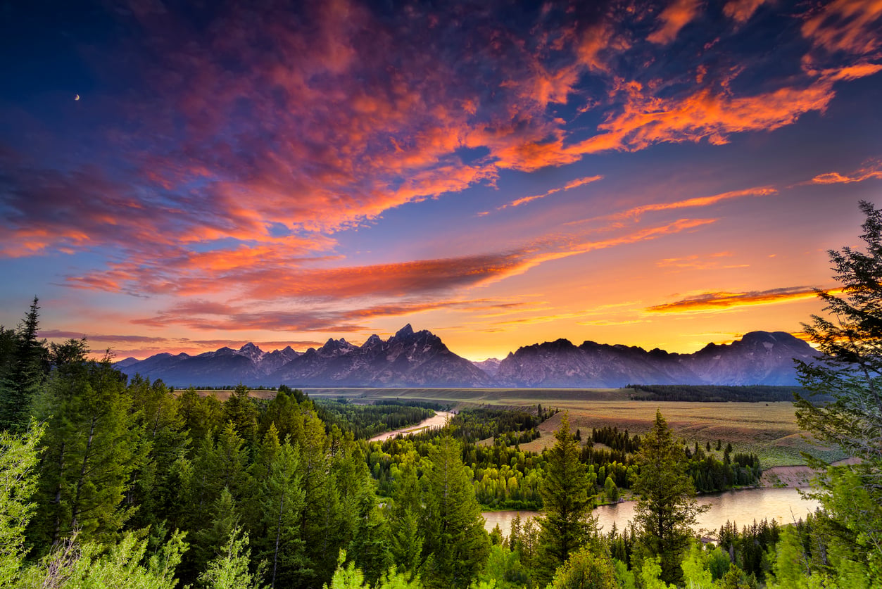 A vibrant sunset over a mountain range with dramatic clouds, a river winding through a lush green forest, and colorful light reflecting on the landscape—one of those American Destinations That Feel Bigger than life.