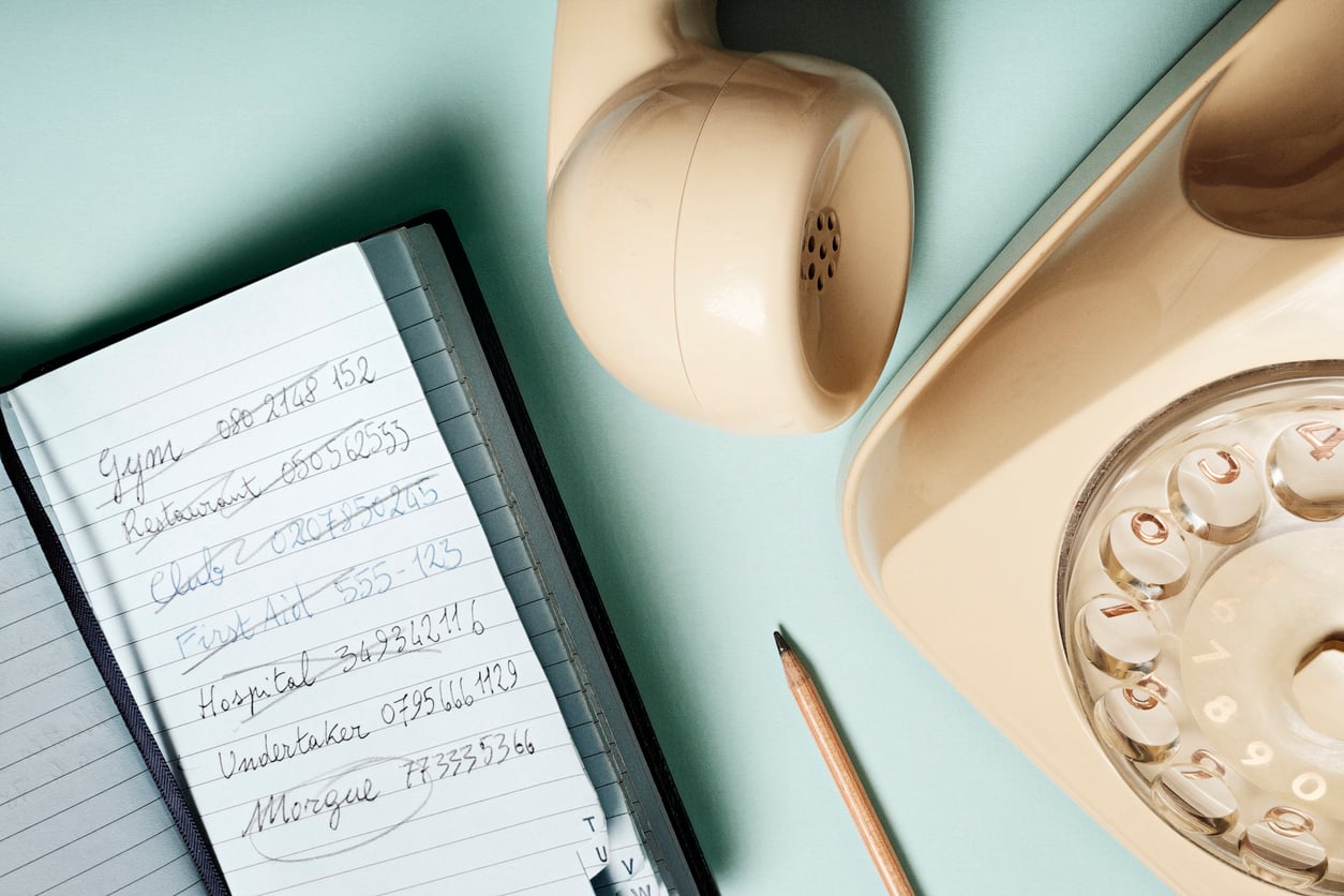 A vintage rotary phone and pencil resting next to an open notebook with a handwritten list of emergency contact numbers.