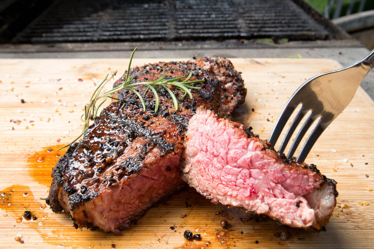 A seared steak topped with a sprig of rosemary on a wooden cutting board, with a fork lifting a sliced piece to reveal a pink, medium-rare center.
