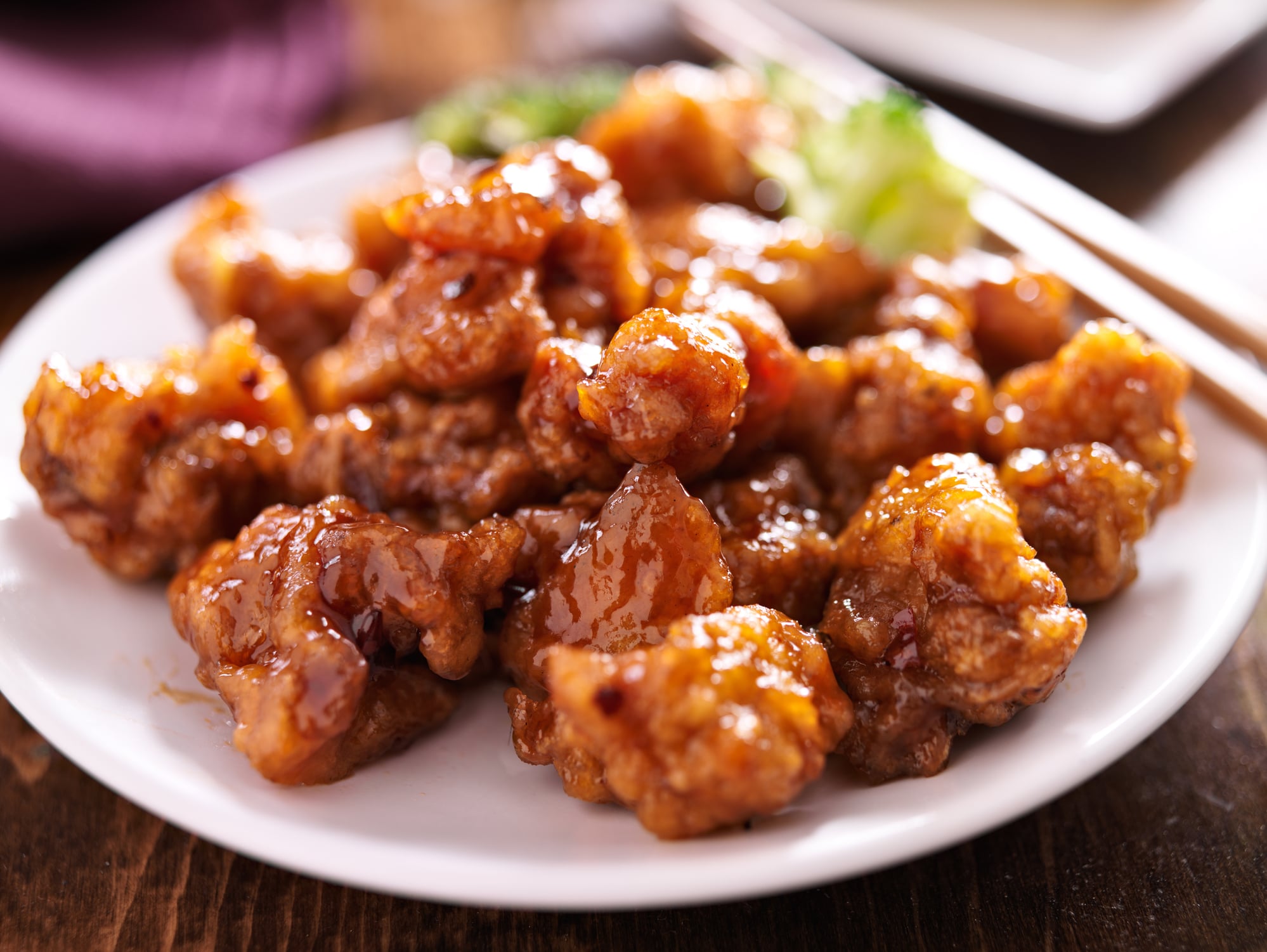 A plate of glazed orange chicken with broccoli florets and wooden chopsticks.