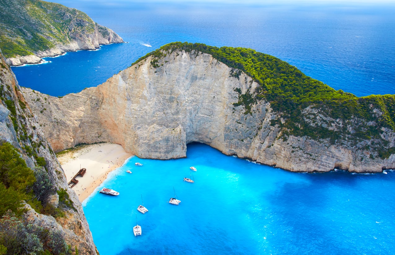 Several boats anchored in the bright blue water of Navagio Beach, surrounded by high limestone cliffs and lush greenery.