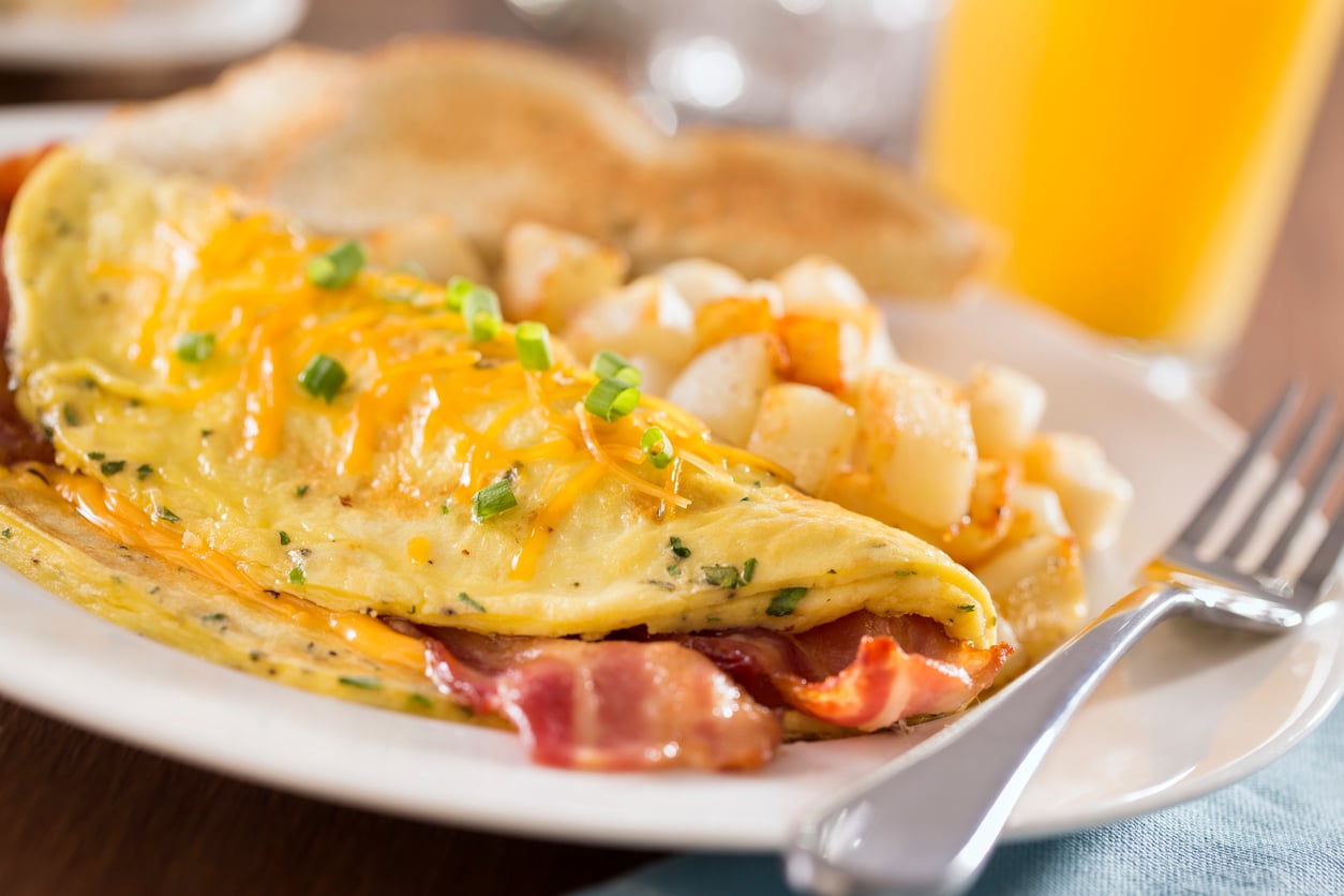 Restaurant orders like cheese and bacon omelet topped with green onions, served with breakfast potatoes and toast, with a fork on the plate and a glass of orange juice nearby.