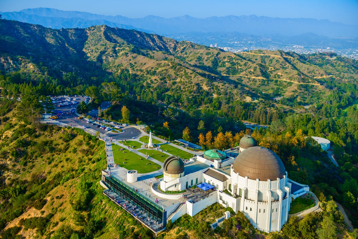 Aerial view of the Griffith Observatory in Los Angeles, situated on a hillside overlooking a city and mountain range.