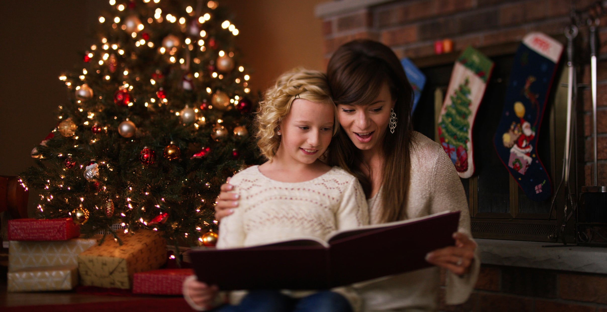 A woman and young girl look at a book together in front of a Christmas tree and a fireplace with hanging stockings.