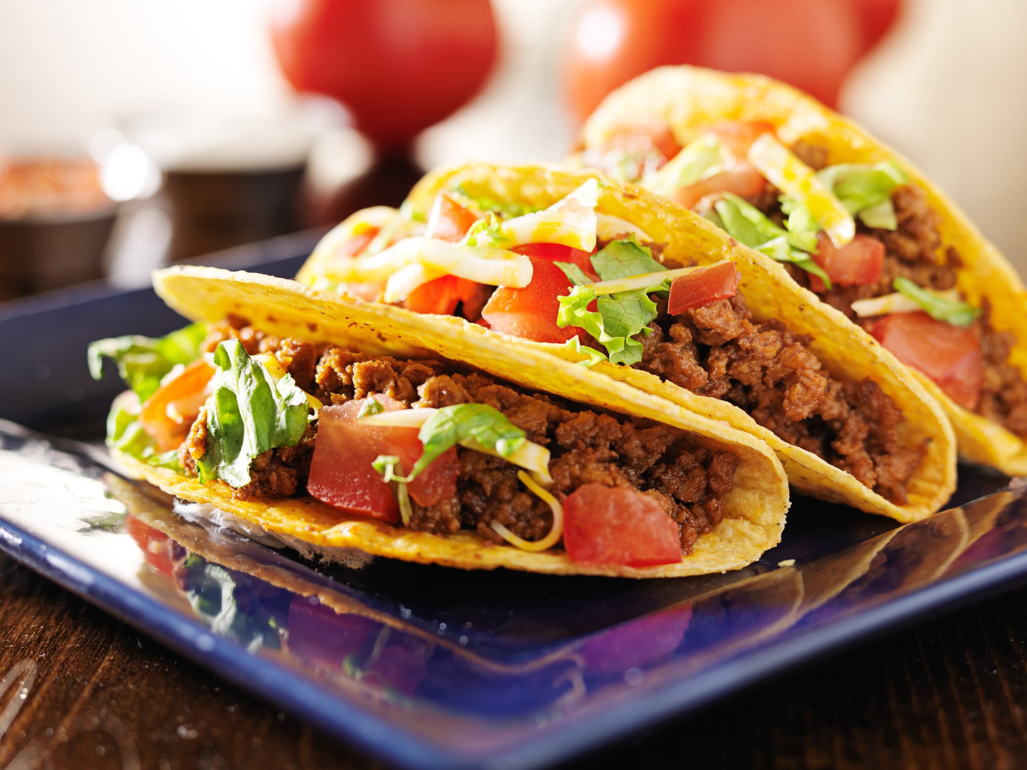 Two hard-shell beef tacos topped with shredded lettuce, diced tomatoes, and cheese on a blue square plate.