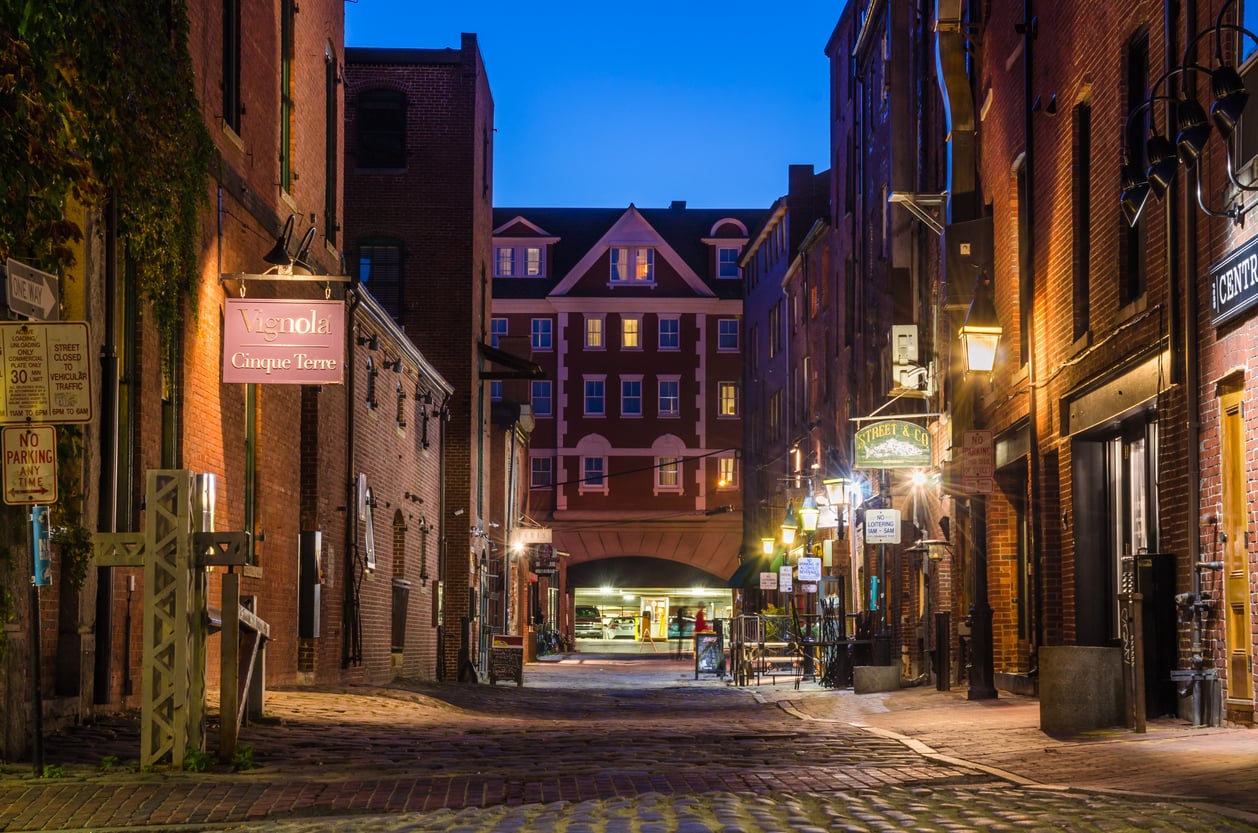 A brick-paved alleyway between historic buildings at twilight, with warm lights illuminating signs for “Vignola Cinque Terre” and “Street & Co.” restaurants.