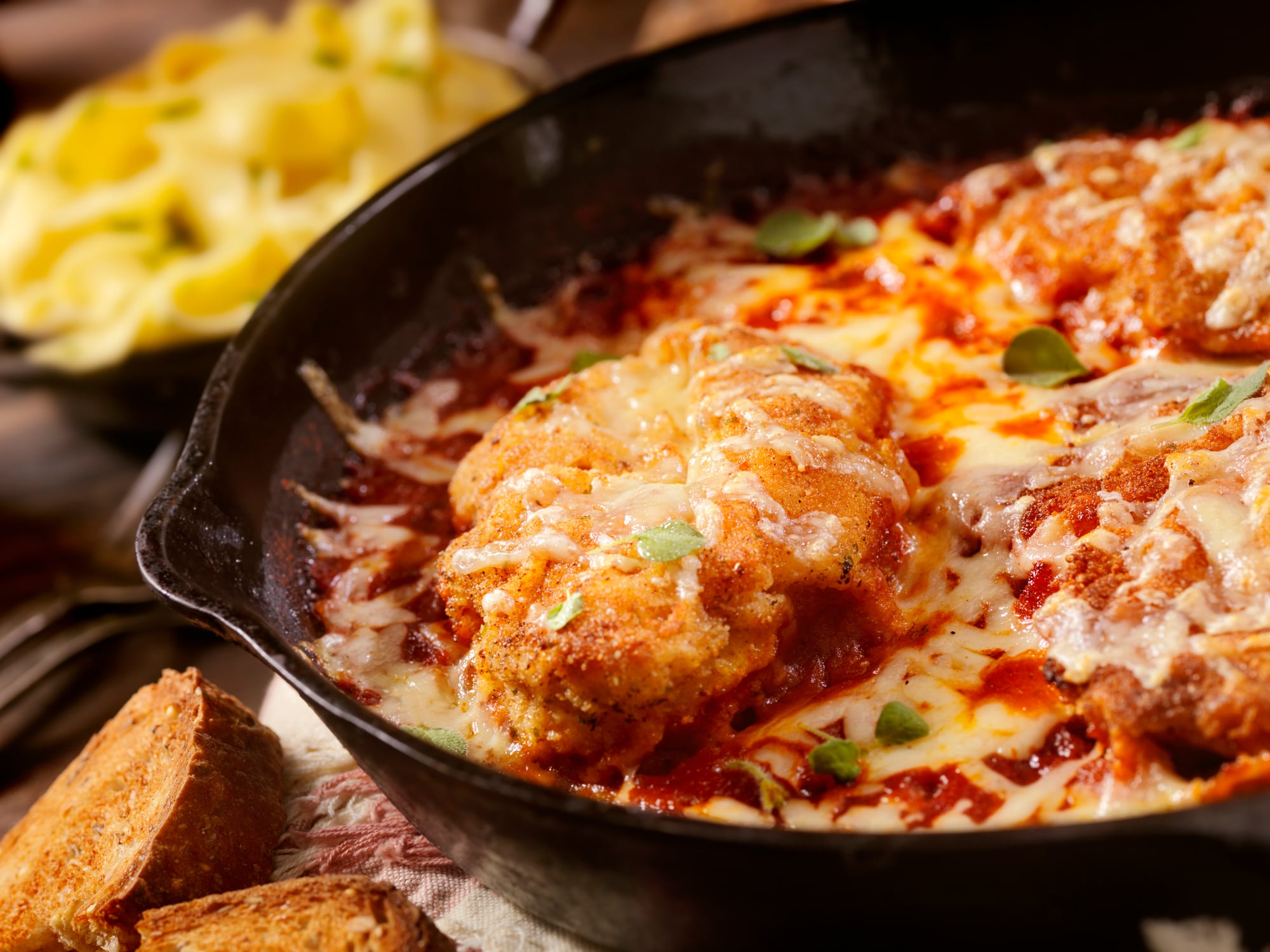 Chicken parmesan in a skillet with melted cheese and red sauce, served with a side of pasta and toasted bread.